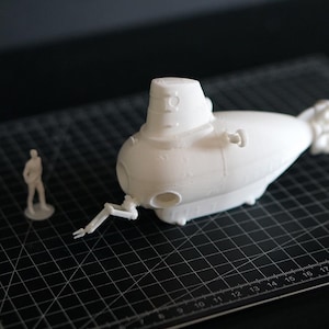 Peut inclure: Maquette de sous-marin blanc imprimée en 3D, avec des détails tels qu'une tour de contrôle, des hélices et des bras articulés. Une petite figurine est à proximité, servant d'échelle. Sur un tapis de coupe noir.
