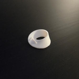 May include: A white, 3D-printed ring-shaped object with a conical design. The object has a smooth surface and a small, protruding tab on one side. The ring is approximately 1 inch in diameter.
