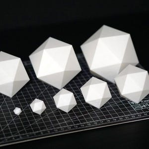 Puede incluir: Una colección de formas geométricas blancas de varios tamaños, que se asemejan a poliedros, están dispuestas sobre una esterilla de corte negra. Las formas tienen múltiples superficies planas y bordes afilados, creando una estética moderna y minimalista.