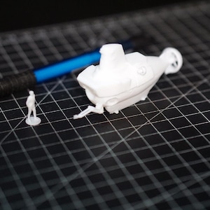Peut inclure: Un modèle de sous-marin blanc imprimé en 3D avec une petite figurine et un stylo bleu sur une surface quadrillée noire. Le sous-marin présente des détails, dont une tour de contrôle et des hélices. La figurine est une représentation miniature d'une personne.