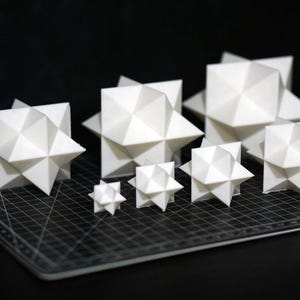 Puede incluir: Una colección de esculturas blancas con forma de estrella geométrica de varios tamaños. Los objetos están dispuestos sobre una esterilla de corte negra, con un fondo oscuro. Las esculturas tienen un diseño facetado.