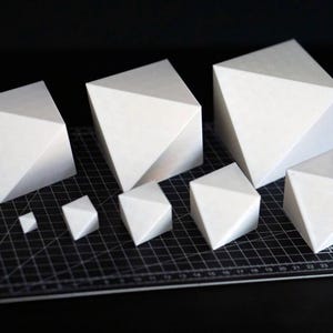 以下が含まれることがあります： 黒いカッティングマットの上に配置された、サイズの異なる8つの白い幾何学的な多面体。マットには白いグリッド線が引かれている。これらの物体は無垢でマットな白色をしており、複数の三角形や四角形の面が組み合わさって複雑な構造を形成している。
