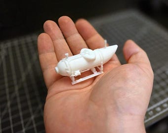 Könnte beinhalten: Ein kleines, weißes Modell-U-Boot mit detaillierten Merkmalen, darunter ein zylindrischer Körper, eine spitze Front und ein kleiner Ständer. Das Modell wird in einer Hand gehalten, um seine Größe und sein kompliziertes Design zu zeigen. Das Modell scheint aus Kunststoff zu sein. Das U-Boot ist etwa 10 cm lang.