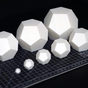 May include: A collection of white geometric shapes of varying sizes, resembling polyhedrons, are arranged on a black cutting mat. The shapes have multiple flat sides and are illuminated to highlight their form. The smallest shape is a sphere.