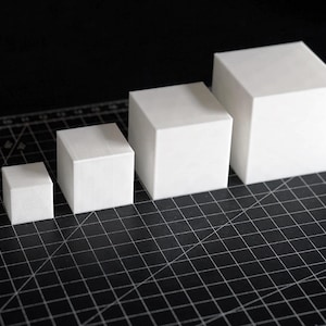 Puede incluir: Cuatro cubos blancos y cuadrados de diferentes tamaños están dispuestos en fila sobre una superficie de cuadrícula negra. Los cubos aumentan de tamaño de izquierda a derecha, mostrando una progresión. Los cubos son sólidos y opacos.