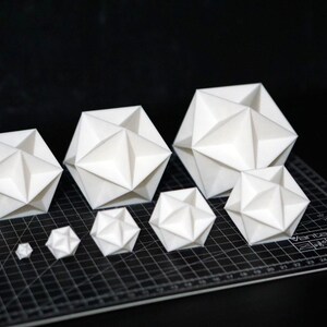 May include: A selection of white geometric paper sculptures in different sizes. The origami-style shapes are displayed on a black cutting mat, creating a visually appealing arrangement. The sculptures have a modern, minimalist aesthetic.