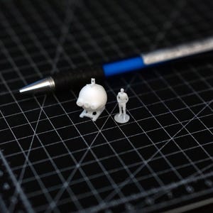 Peut inclure: Un modèle de crâne blanc imprimé en 3D et une figurine humaine miniature sont présentés sur un tapis de découpe noir. Un stylo noir avec une pointe argentée et des accents bleus est en arrière-plan. Le crâne mesure environ 2,5 cm de haut.