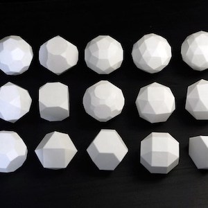 Könnte beinhalten: Anordnung von fünfzehn weißen geometrischen Formen mit verschiedenen facettierten Designs. Die Objekte, darunter Kugeln und Polyeder, sind auf einer schwarzen Oberfläche angeordnet, wobei die Beleuchtung die Winkelflächen hervorhebt.