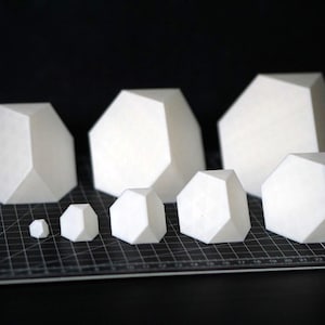 May include: A selection of white, geometric paper sculptures in various sizes. The shapes are multi-faceted, resembling stylized rocks or crystals. They are arranged on a black cutting mat, creating a contrast with the white objects.
