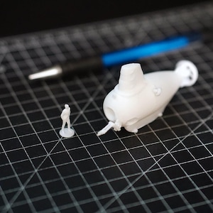 Peut inclure: Un modèle de sous-marin miniature blanc avec une petite figurine à côté. Le sous-marin a un design détaillé, comprenant une hélice et une tour de contrôle. Un stylo est en arrière-plan. Les objets sont sur une surface de grille noire.