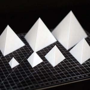 Puede incluir: Una colección de pirámides de papel blanco de varios tamaños dispuestas sobre una esterilla de corte negra. Las pirámides varían de pequeñas a grandes, mostrando un diseño geométrico. La esterilla tiene un patrón de cuadrícula y un logotipo de Vantage.