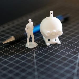 Peut inclure: Une figurine blanche imprimée en 3D représente une personne à côté d'un objet sphérique avec une petite antenne. L'objet a trois éléments circulaires. Un stylo noir avec une pointe argentée est en arrière-plan sur une surface quadrillée.