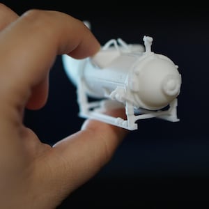 Peut inclure: Un petit modèle blanc d'un objet cylindrique avec des détails, notamment des protubérances et des supports. Le modèle est tenu entre les doigts sur un fond sombre, soulignant son design et sa structure complexes.