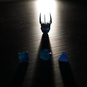May include: A dramatic image of a blue crown and three blue dice, all casting shadows. The crown, lit from above, is the focal point. The dice are arranged below. The scene is set against a dark background, creating a sense of mystery.