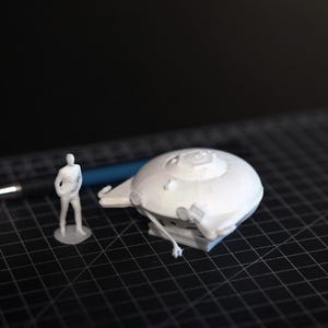 Peut inclure: Modèle blanc imprimé en 3D d'un vaisseau spatial avec une petite figurine humaine et un stylo sur une surface de grille noire. Le vaisseau spatial a un dessus arrondi et plusieurs éléments en saillie.