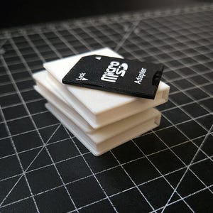 Puede incluir: Una tarjeta adaptadora Micro SD negra con texto blanco descansa sobre una pila de tres bloques rectangulares blancos. La tarjeta adaptadora dice "Micro SD Adapter" y tiene un símbolo de bloqueo. Los elementos están sobre una superficie negra con un patrón de cuadrícula.