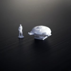 Peut inclure: Maquette blanche d'un vaisseau spatial et d'une figurine. Le vaisseau spatial a un dessus arrondi avec des détails et repose sur une base carrée. La figurine est une forme humaine simple, debout à côté du vaisseau spatial.