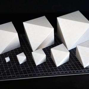 Peut inclure: Ensemble de sculptures géométriques blanches de différentes tailles. Chaque sculpture présente une face triangulaire et une forme cubique. Les objets sont disposés sur un tapis de coupe noir, créant un contraste.