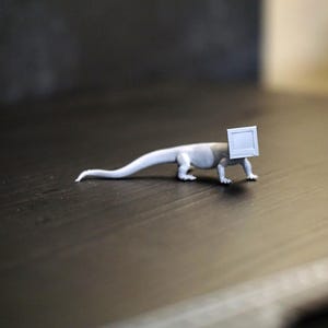 Puede incluir: Una pequeña figura de lagarto gris con una cabeza cuadrada, sobre un fondo oscuro. El lagarto está en posición de caminar, con la cola extendida. La cabeza es un cuadrado blanco, lo que le da al lagarto una apariencia única.