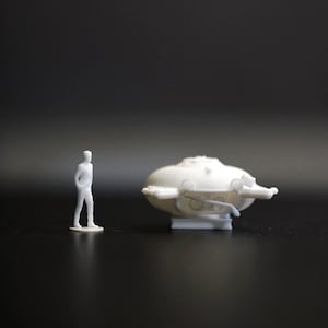 Peut inclure: Un modèle miniature blanc d'un sous-marin futuriste avec une petite figurine d'une personne debout à côté. Le sous-marin a un corps arrondi avec des structures en forme d'ailes et un avant saillant. La figurine se tient debout, les mains dans les poches.