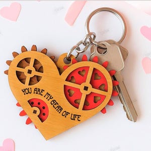 Könnte beinhalten: Holz-Schlüsselanhänger in Herzform mit Zahnrad-Design und dem Text "YOU ARE MY GEAR OF LIFE". Der Schlüsselanhänger hat ein rot-braunes Farbschema und ist an einem Schlüsselring mit Schlüsseln befestigt. Die Materialbreite beträgt 3 mm.