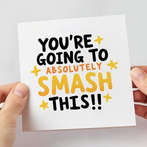 May include: A white greeting card with the text "YOU'RE GOING TO ABSOLUTELY SMASH THIS!!" in black and yellow lettering. The card is held by two hands.
