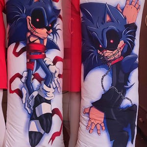 May include: A white rectangular body pillow case featuring a graphic illustration of a dark blue hedgehog character with red glowing eyes, sharp teeth, and red bandages on the left side, and a chained character with a metal muzzle on the right side.
