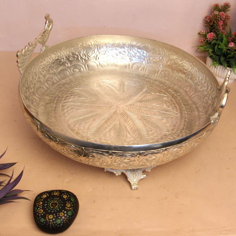 May include: Silver-colored decorative bowl with ornate floral patterns and handles. The bowl has a detailed, embossed design on the interior and exterior. It is supported by three small feet and is likely used for display or serving.
