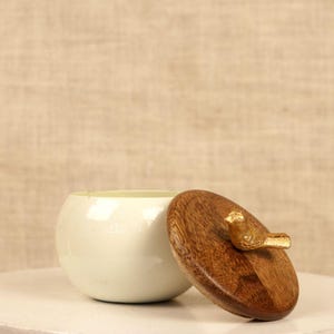 May include: A small, off-white ceramic bowl with a wooden lid. The lid is oval-shaped and features a small, gold-colored bird as a handle. The bowl is partially open, revealing its interior. The background is a neutral beige.