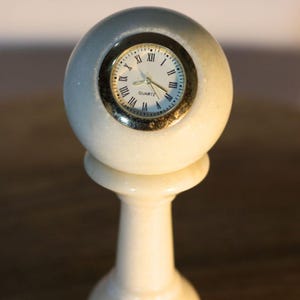 May include: A white marble quartz clock with a round face and Roman numerals. The clock is set in a spherical marble casing and sits atop a pedestal base. The clock's hands indicate the time.