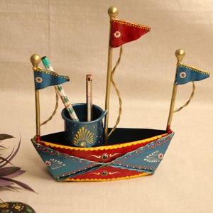 May include: A decorative metal desk organizer shaped like a boat, painted in blue, red, and gold. It features three small flags and a central cup for pens and pencils. The boat is adorned with floral patterns.