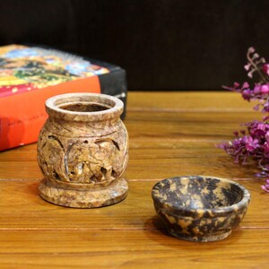 Peut inclure: Un vase en pierre sculptée et un petit bol. Le vase est cylindrique avec une large ouverture et un relief détaillé d'éléphants. Le bol est rond avec un motif tacheté. Les deux articles sont dans des tons de brun et de beige.