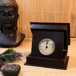 May include: A dark brown wooden clock with a silver-colored clock face. The clock face has Roman numerals and the words "Imperial India" printed on it. The clock is in an open box-like frame.