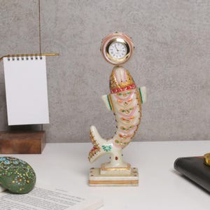 May include: A decorative fish-shaped clock made of white marble, adorned with gold, red, and pink accents. The clock face is round with a gold frame. The fish has detailed scales and fins, and sits on a rectangular base.