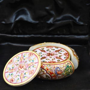 May include: A set of white marble coasters with intricate floral and paisley designs in red, gold, and green. The coasters are round with a gold trim and are housed in a matching holder, showcasing a peacock design.