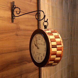May include: A double-sided wooden clock with a checkerboard pattern on the sides, suspended from a black metal bracket. The clock face is cream-colored with black numerals and hands. The clock is mounted on a wooden wall.