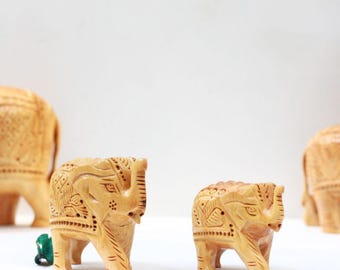 May include: Wooden elephant figurines with intricate carvings. The elephants are light brown and have detailed patterns on their bodies. The image shows multiple elephants of varying sizes, suggesting a family or set of decorative items.