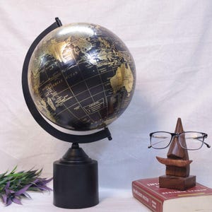 May include: A black and gold decorative globe with a black base and stand. The globe features gold continents and a black ocean. The globe is a decorative item, suitable for a desk or shelf. A wooden sculpture of a face holds a pair of glasses.