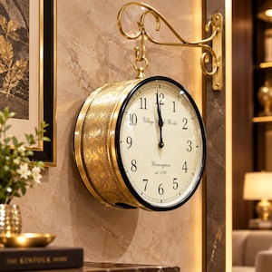 May include: A double-sided wall clock with a gold-toned metal frame and a cream-colored face. The clock features black Arabic numerals, black hands, and the text Village Clock Works Birmingham est. 1792. It hangs from a decorative gold metal bracket.
