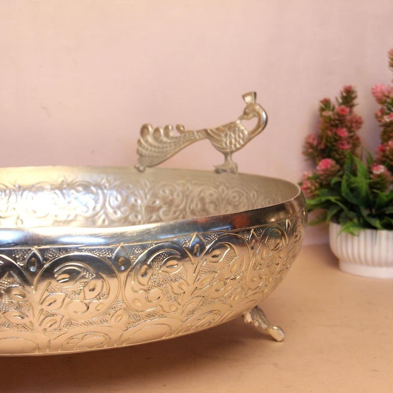 May include: Silver-colored decorative bowl with a detailed floral pattern and a peacock-shaped handle. The bowl has three small feet and is designed for decorative purposes. The bowl is made of metal and has a shiny finish.