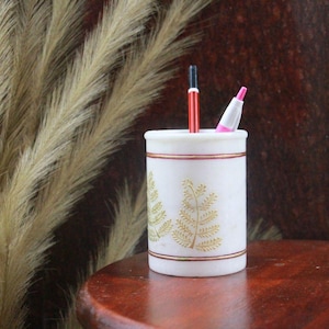 May include: A white marble pencil holder with gold leaf design and red and gold trim. The holder contains a red and black pencil and a pink highlighter. The pencil holder is on a wooden table.