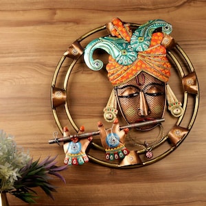 May include: A decorative metal wall hanging featuring a face with a turban and flute. The face is bronze-colored with closed eyes and a red dot on the forehead. The turban is orange and green with intricate patterns. The hands are holding a flute.