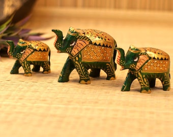 May include: Three wooden elephant figurines. The elephants are painted green with gold and red accents. The figurines are standing on a light brown surface. The elephants are of varying sizes, with the largest in the center.