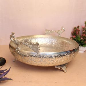 May include: Silver-colored decorative bowl with peacock accents. The bowl has a detailed floral pattern and is supported by three small feet. Two peacock figures are on the rim. The bowl is likely made of metal and is suitable for decorative purposes.