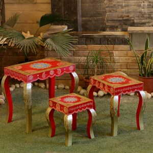 Puede incluir: Conjunto de tres mesas decorativas con tapas rojas y patas doradas. Las mesas tienen un borde festoneado y un diseño floral detallado en la tapa. Las patas son curvas y pintadas de dorado.