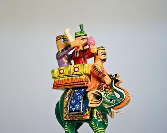 May include: A hand-painted wooden elephant figurine carrying three figures. The elephant is green with orange and yellow accents. The figures are dressed in colorful clothing, with one holding a pink flower. The figurine has a decorative platform.
