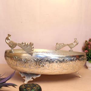 May include: Silver-colored decorative bowl with peacock handles and ornate floral patterns. The bowl is oval-shaped and rests on three decorative feet. The handles are detailed with peacock designs. A stylish piece for home decor.