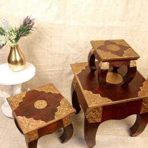 May include: Three wooden accent tables with dark brown tops and ornate gold-colored metal trim. The tables have curved legs and are of varying sizes, with the smallest table resting on the largest. A gold vase with flowers is visible in the background.