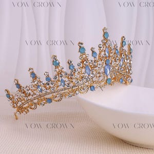 Op de afbeelding: Een goudkleurige kroon met blauwe en heldere edelstenen. De kroon heeft een gedetailleerd ontwerp met meerdere punten en wordt gepresenteerd op een wit oppervlak. De tekst "VOW CROWN" is zichtbaar op de achtergrond.