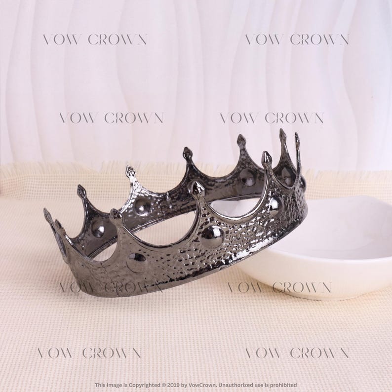 Op de afbeelding: Een donkere metalen kroon met een gehamerde textuur en een reeks puntige bogen. Het oppervlak heeft een reflecterende afwerking met cirkelvormige accenten aan de basis van elk punt. De tekst VOW CROWN is herhaald op de achtergrond.